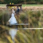 NZ Wedding Photographer photo of bride and groom stand holding hands and looking at each other shot through long grass across a pond